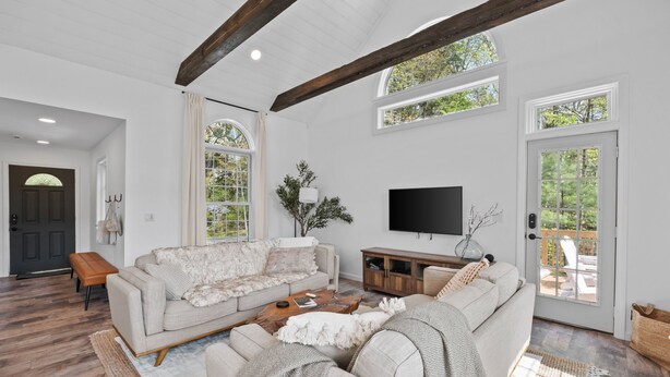 A spacious living room with a large sectional sofa, a coffee table, and a flat-screen TV. There are large windows and a door leading to an outdoor patio. - kids secret room, Canoes/Kayaks, fire pit, ev charger. (Center Harbor)