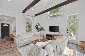 A spacious living room with a large sectional sofa, a coffee table, and a flat-screen TV. There are large windows and a door leading to an outdoor patio. - kids secret room, Canoes/Kayaks, fire pit, ev charger. (Center Harbor)