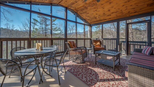 A covered outdoor patio with a wooden ceiling, large windows, wicker furniture, and a view of the surrounding landscape. - Cozy Tucked Away Log Cabin- Lake/Mountain views! (Lake Lure)