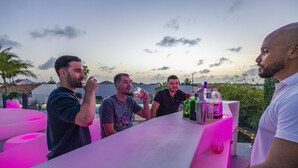 A bar area with a white countertop and various bottles, set against a backdrop of a sunset sky and palm trees. - Villa Cocolishi:Luxe Pool Retreat 5 Min->Beaches (Noord)