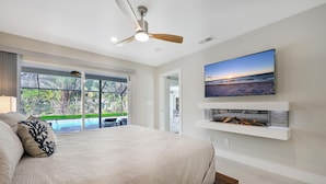 A bedroom with a large bed, a ceiling fan, a TV mounted above a fireplace, and a view of a pool and greenery through a sliding glass door. - PROMO 5% WEEKLY AND 10% MONTHLY DISCOUNT
Tropical Dream House with Pool (Naples)