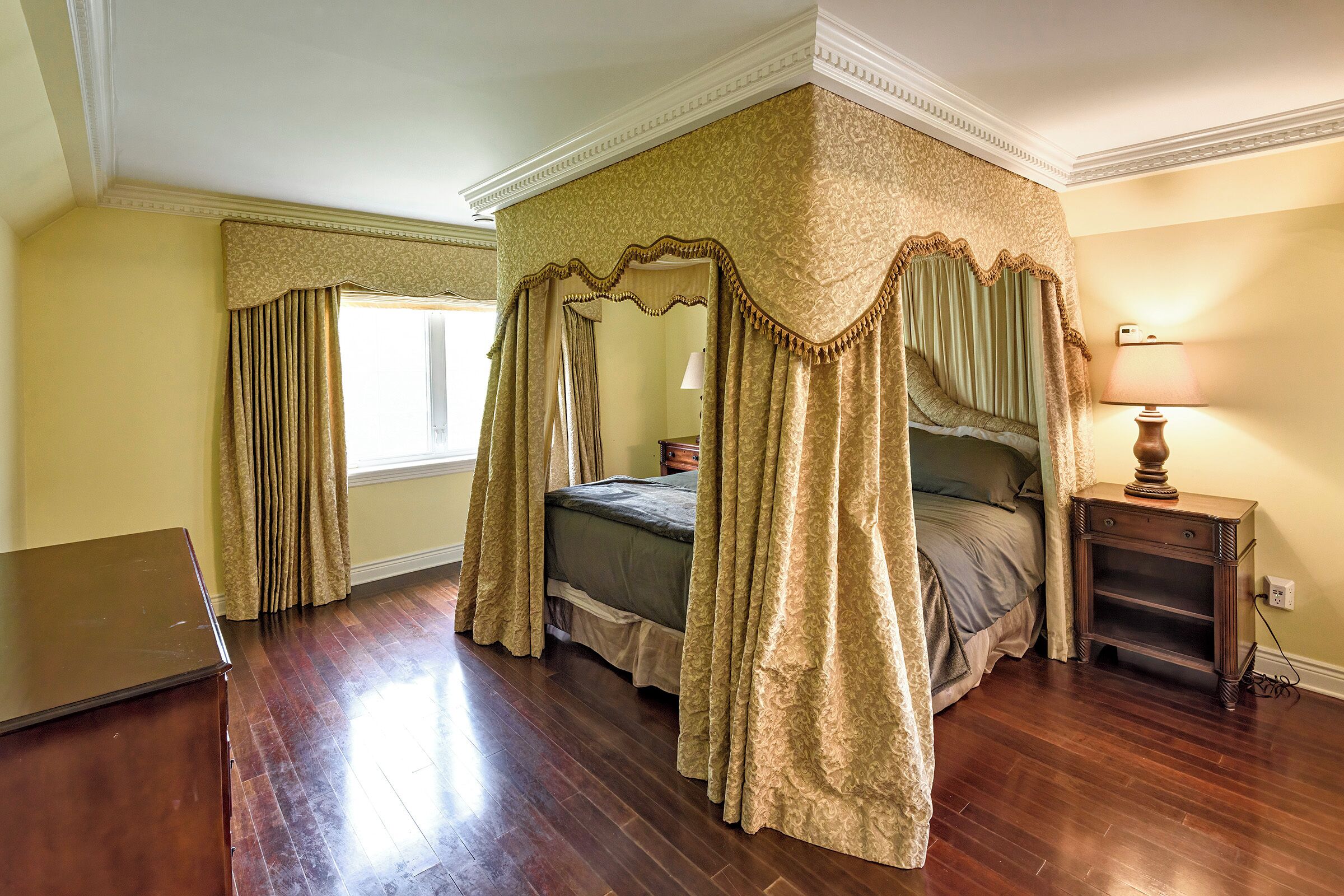 A four-poster canopy bed with curtains, a bedside table with a lamp, and a laptop on a wooden dresser.