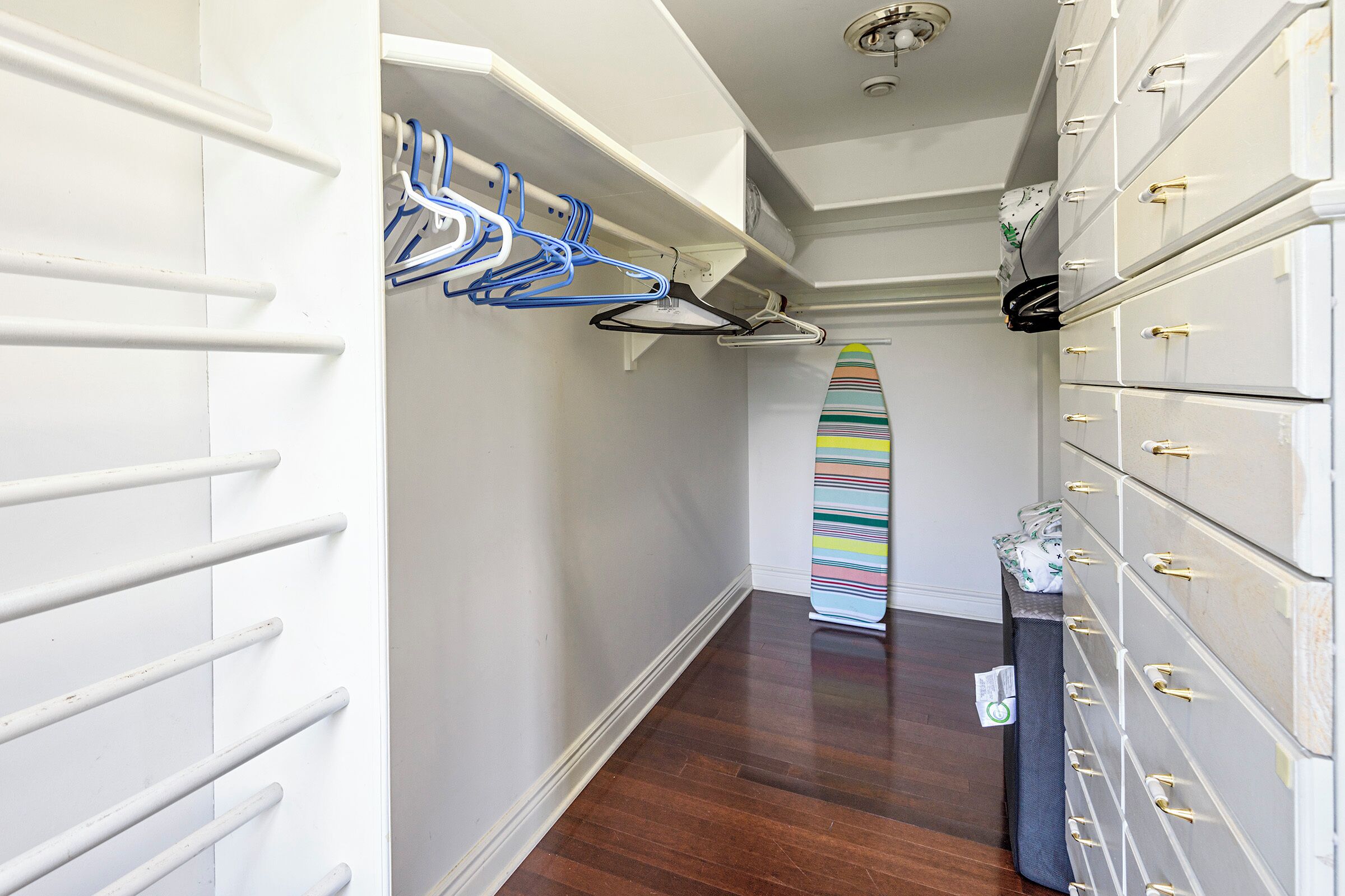 A hallway with a white wall, a wooden floor, a surfboard leaning against the wall, and a set of white storage drawers with gold handles.