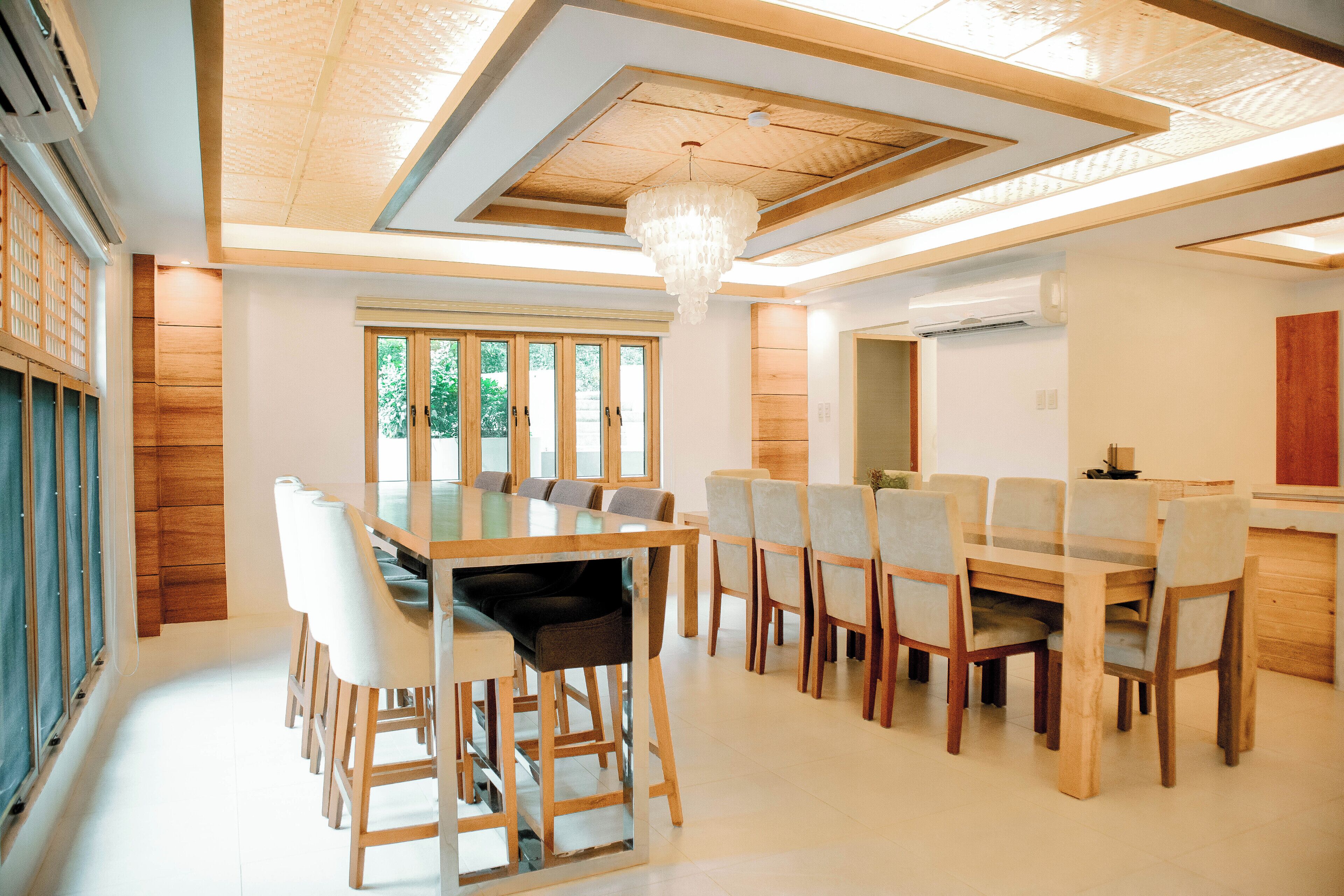 A spacious dining area with a large wooden table and chairs, a chandelier, and a view of greenery through the windows.