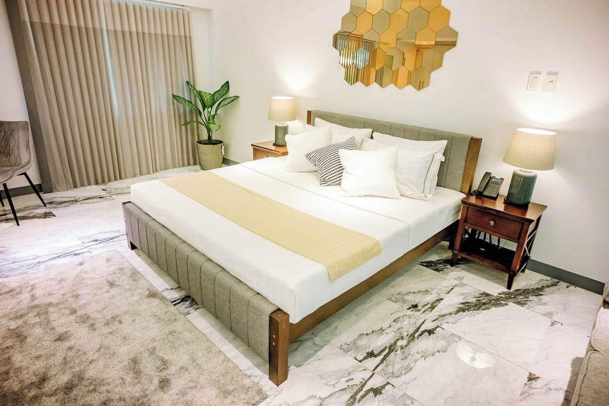 A hotel room with a large bed, two bedside tables with lamps, a potted plant, and a decorative wall piece.