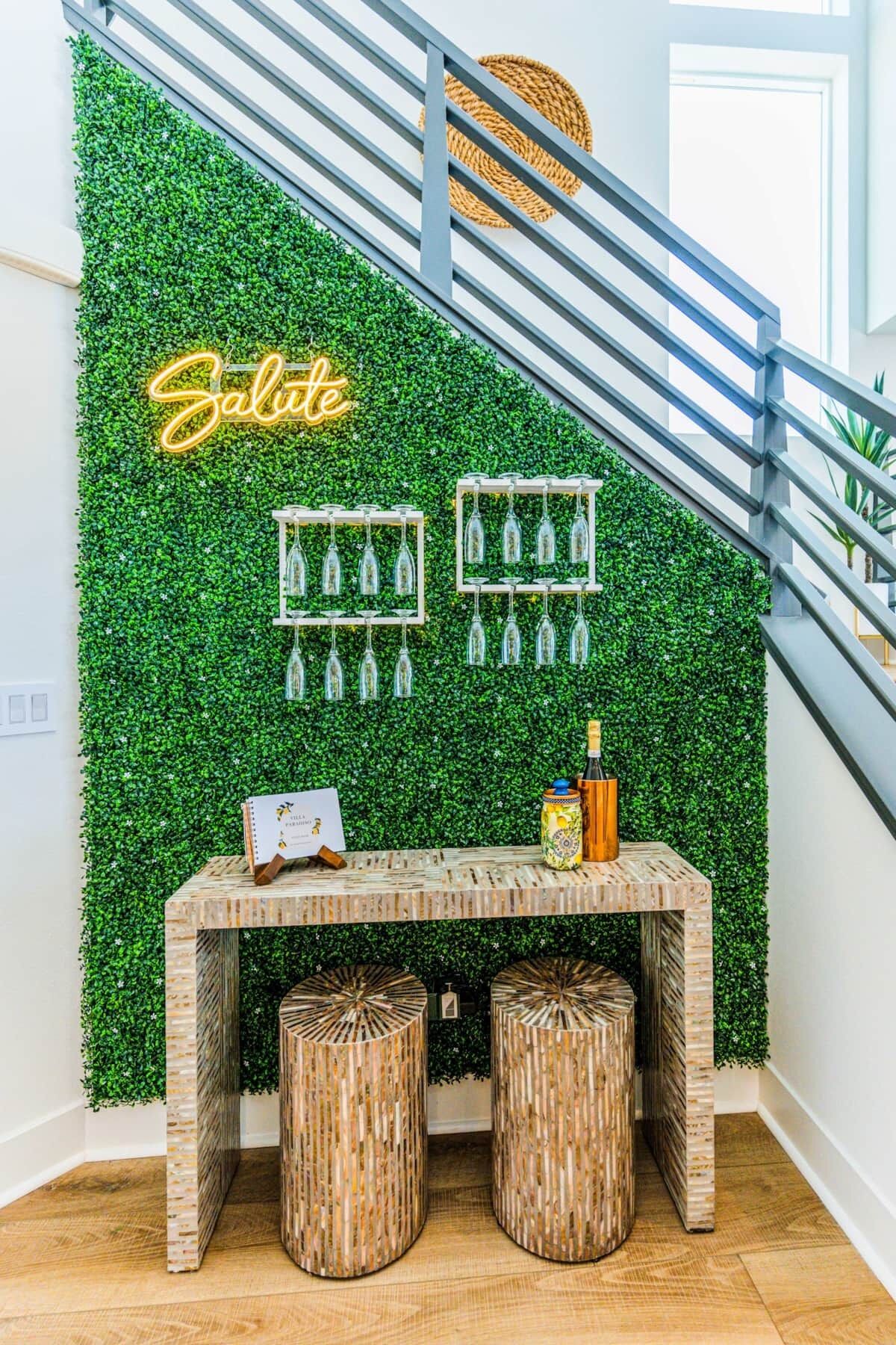 Una escalera con una pared verde, un letrero de neón, copas de vino y una pequeña mesa con una botella.