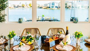 A dining table set for two with wicker chairs, a fruit platter, and a bottle of champagne. - 4 Private EnSuites, 270* Lake View, pool+spa, Sleeps 12 (Indio)