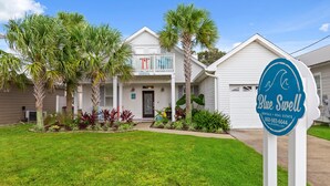 Une maison à deux étages avec un porche à l’avant, des palmiers et une enseigne indiquant « Blue Swell Rentals & Real Estate ».