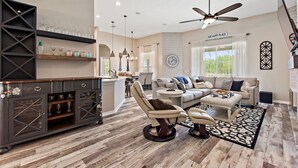 A spacious living room with a high ceiling, a ceiling fan, a fireplace, and a dining area with a kitchen in the background. - Serenity Now By Shine Villas At Watersong #416 5 Bedroom Villa (Davenport)