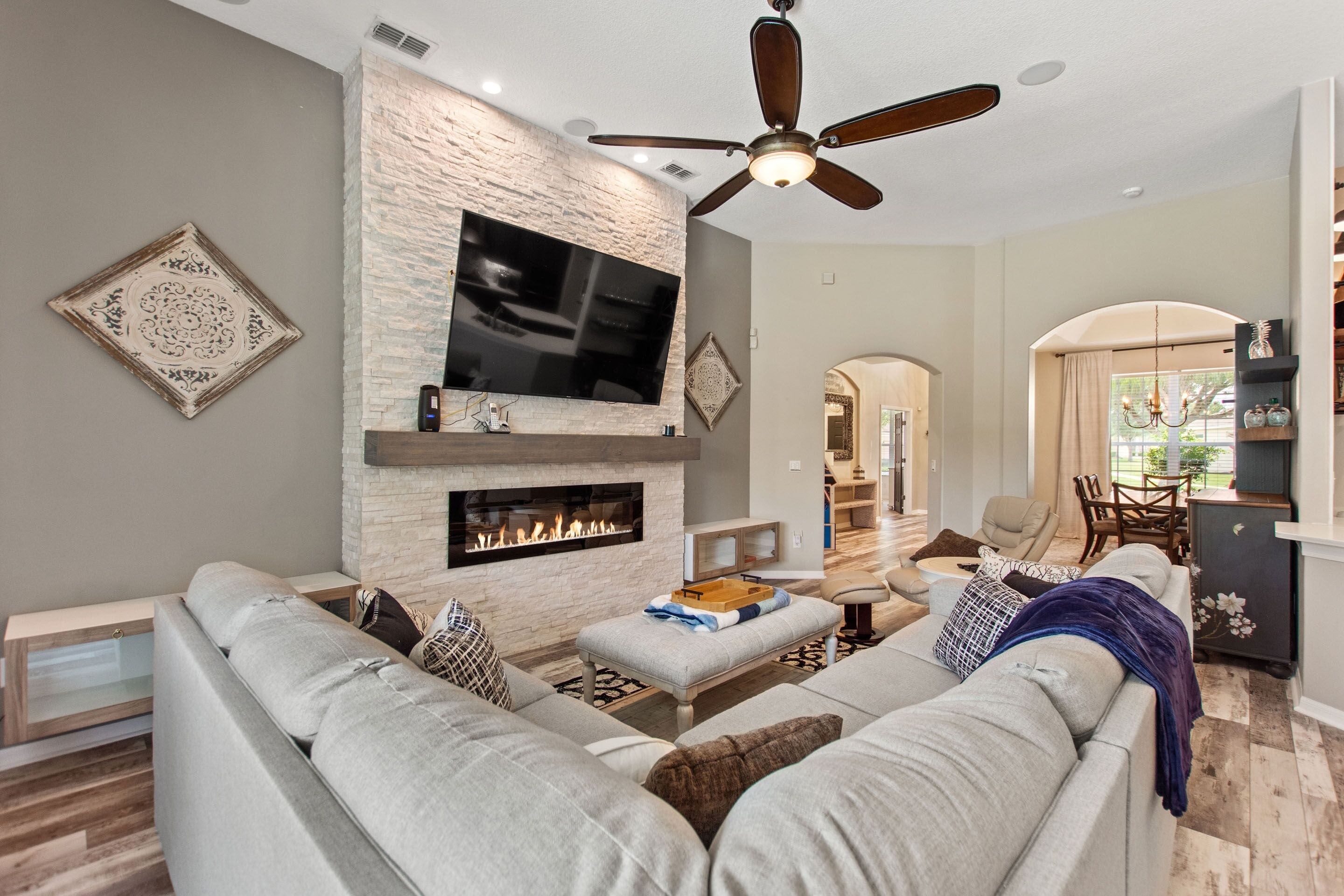 A modern living room with a stone fireplace, a flat-screen TV, and a ceiling fan.