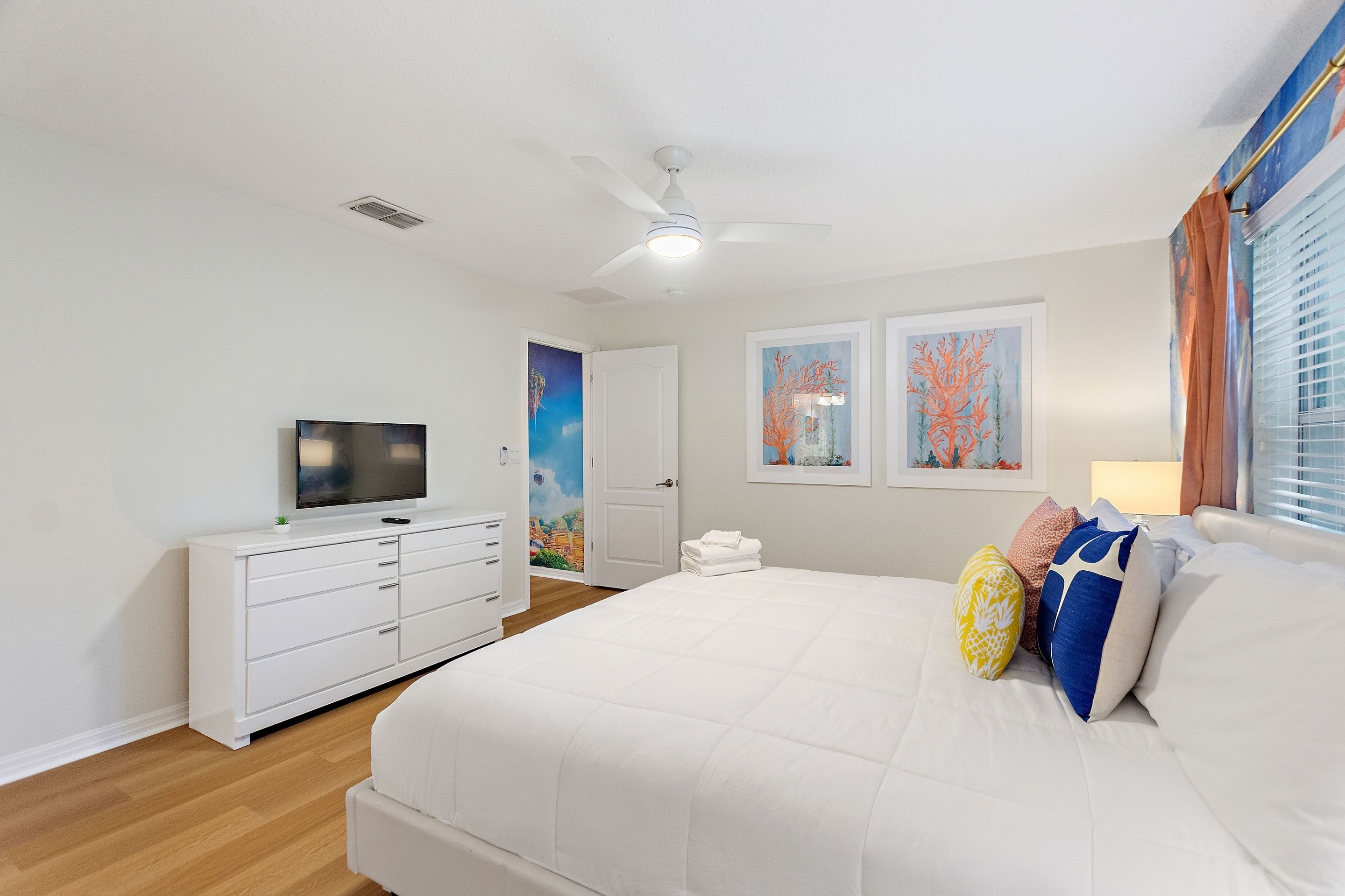 Un dormitorio con una cama, un televisor sobre un tocador blanco y dos obras de arte enmarcadas en la pared.