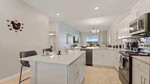 A modern kitchen with white cabinetry, a central island, and a dining area with chairs. - Edgewater Escape At Storey Lake By Shine Villas 2 Bedroom Apts (Kissimmee)