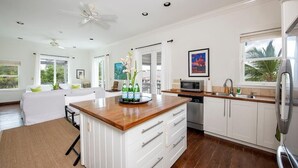 A modern kitchen with white cabinetry, a wooden countertop, and a central island. There is a dining area with a white sofa and a ceiling fan. - Sand Dollar Cottage 2 Bedroom Cottage (Rock Sound)