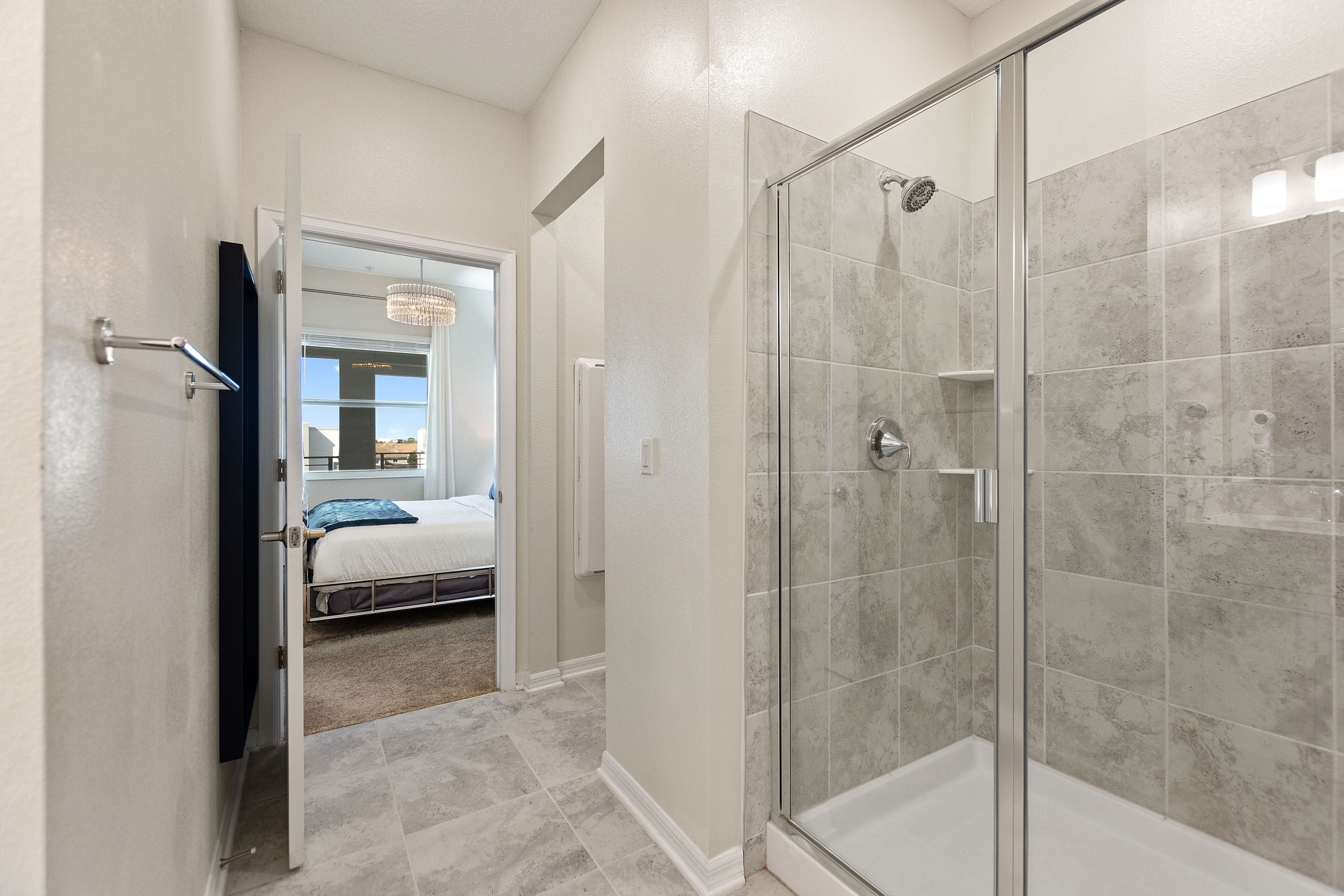 Une salle de bain moderne dotée d’une douche à parois de verre, d’un pommeau de douche apparent et d’une porte donnant sur une chambre à coucher avec un lit et un lustre.