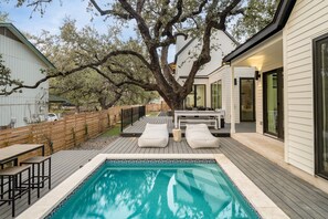 A modern outdoor pool area with a wooden deck, lounge chairs, and a large tree.