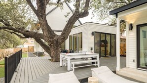 A modern outdoor deck with a wooden floor, white furniture, and a large tree. - 6BDR Luxe Pool Retreat • City Views, Ping Pong, Foosball, Guest House | Henley (Austin)