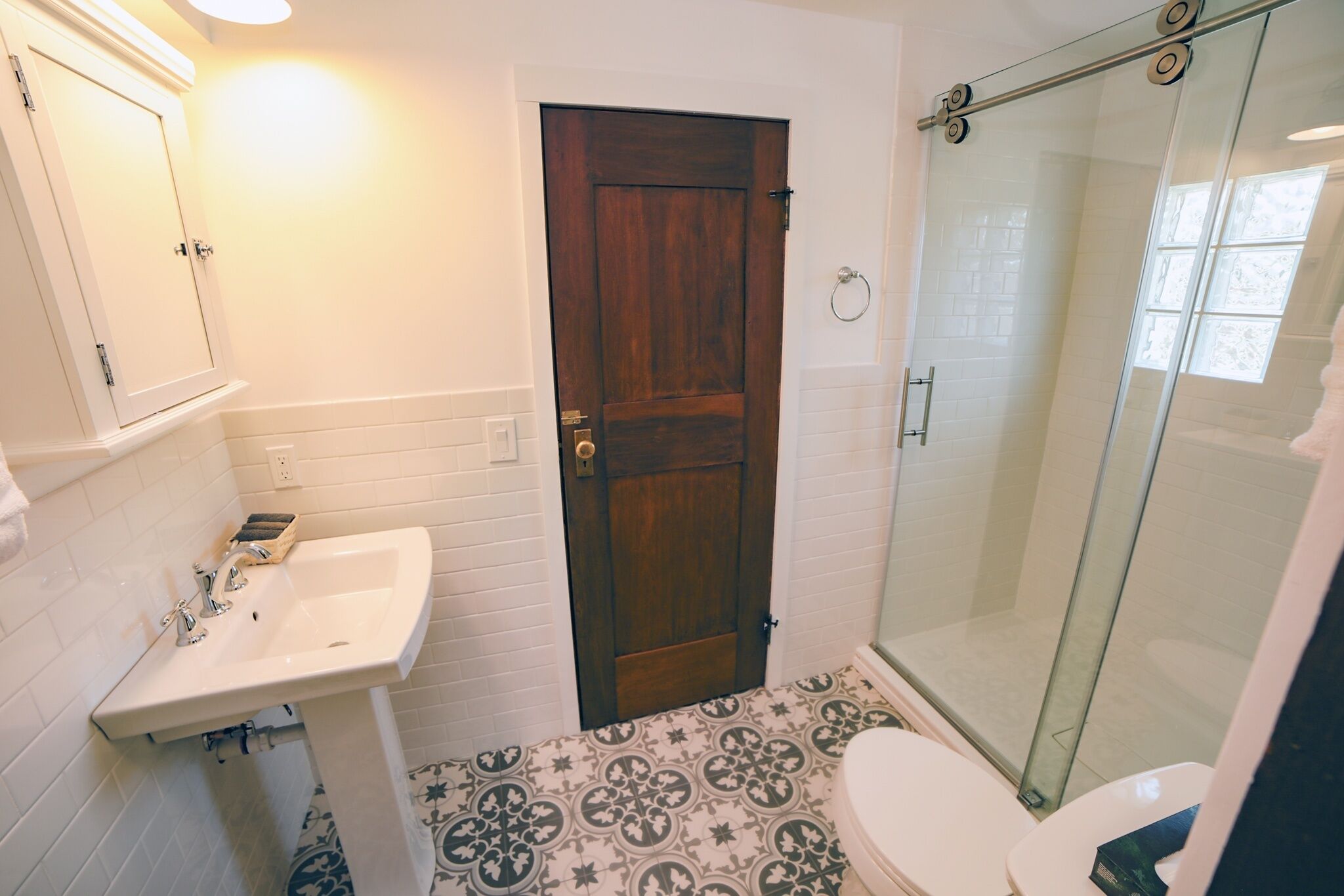 Un baño con lavabo blanco, puerta de madera, mampara de cristal y baldosas de suelo estampadas.