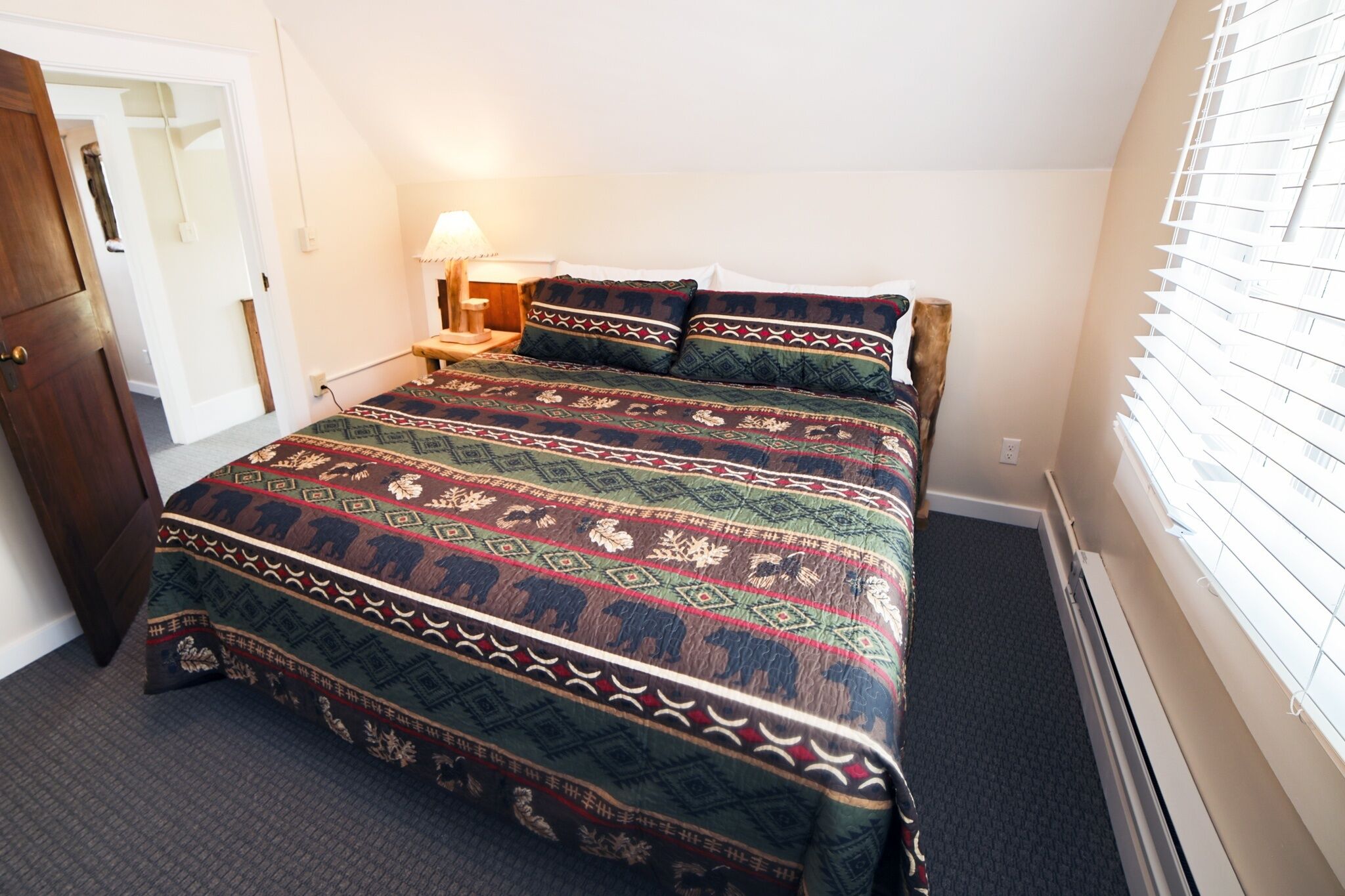 A bedroom with a large bed, a nightstand with a lamp, a wooden door, and a window with blinds.