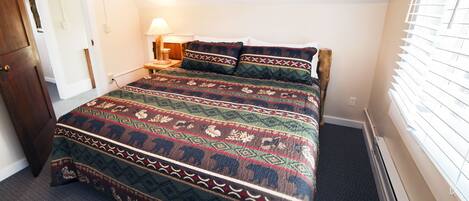 A bedroom with a large bed, a nightstand with a lamp, a wooden door, and a window with blinds.