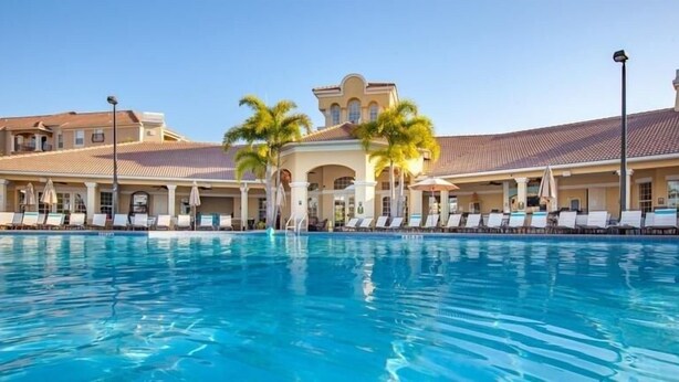A large swimming pool with lounge chairs and umbrellas, surrounded by a building with a tiled roof and palm trees. - Vista Cay Resort, Next To Universal 821 3 Bedroom Townhouse (Orlando)