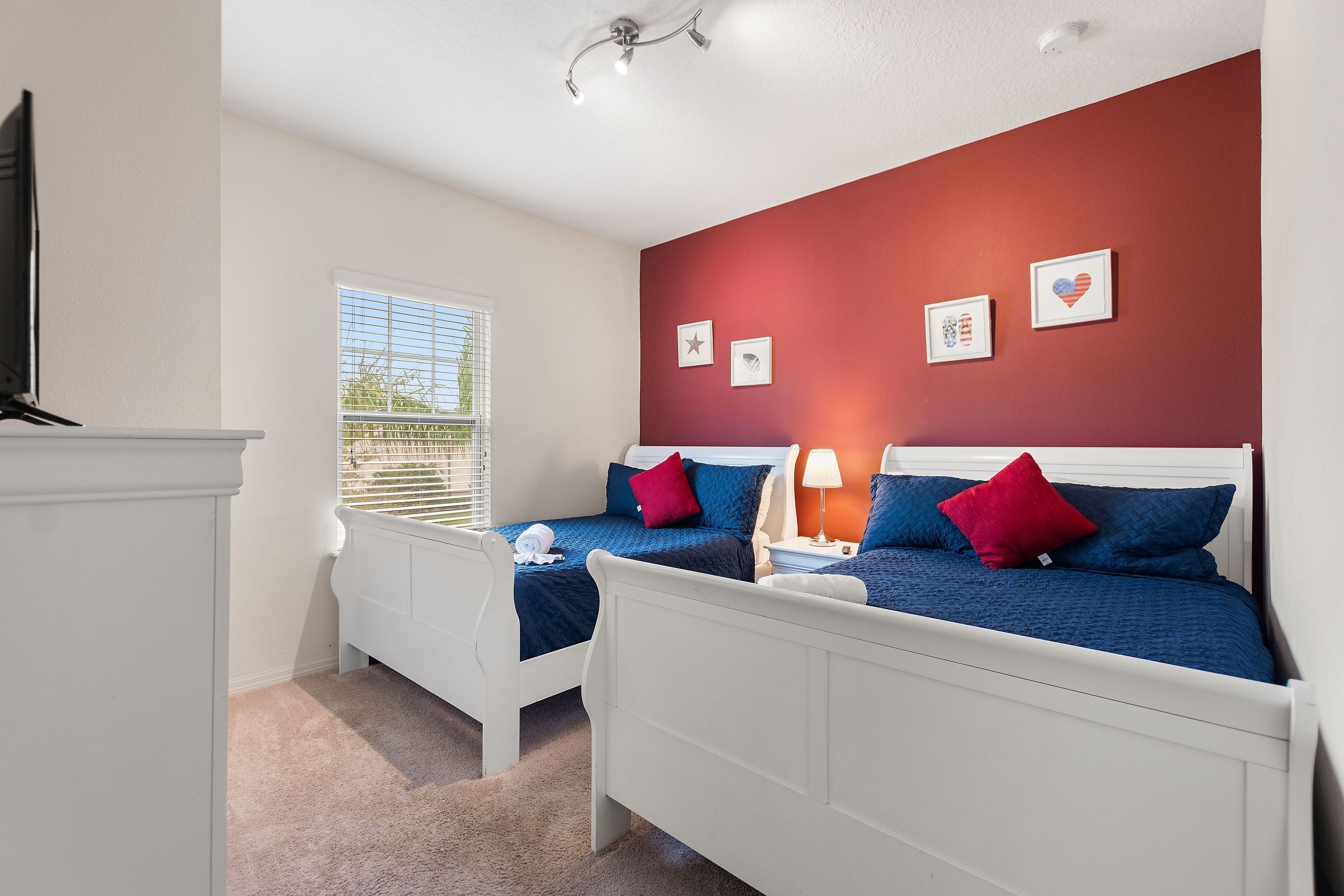 A bedroom with two beds, white headboards, a window with blinds, and a red accent wall with framed pictures.