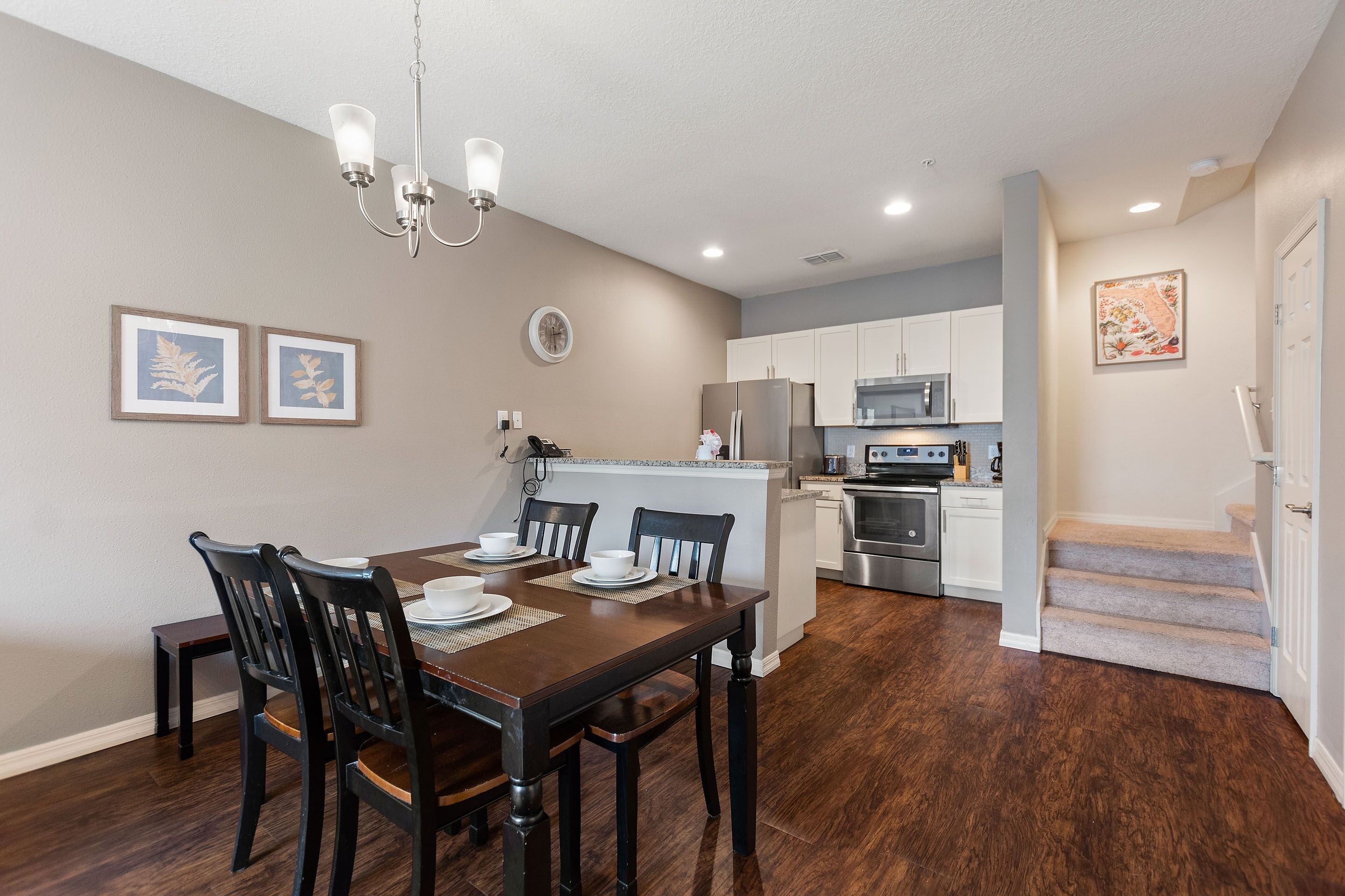 A dining area with a wooden table and chairs, a chandelier, framed artwork on the wall, and a kitchen in the background.