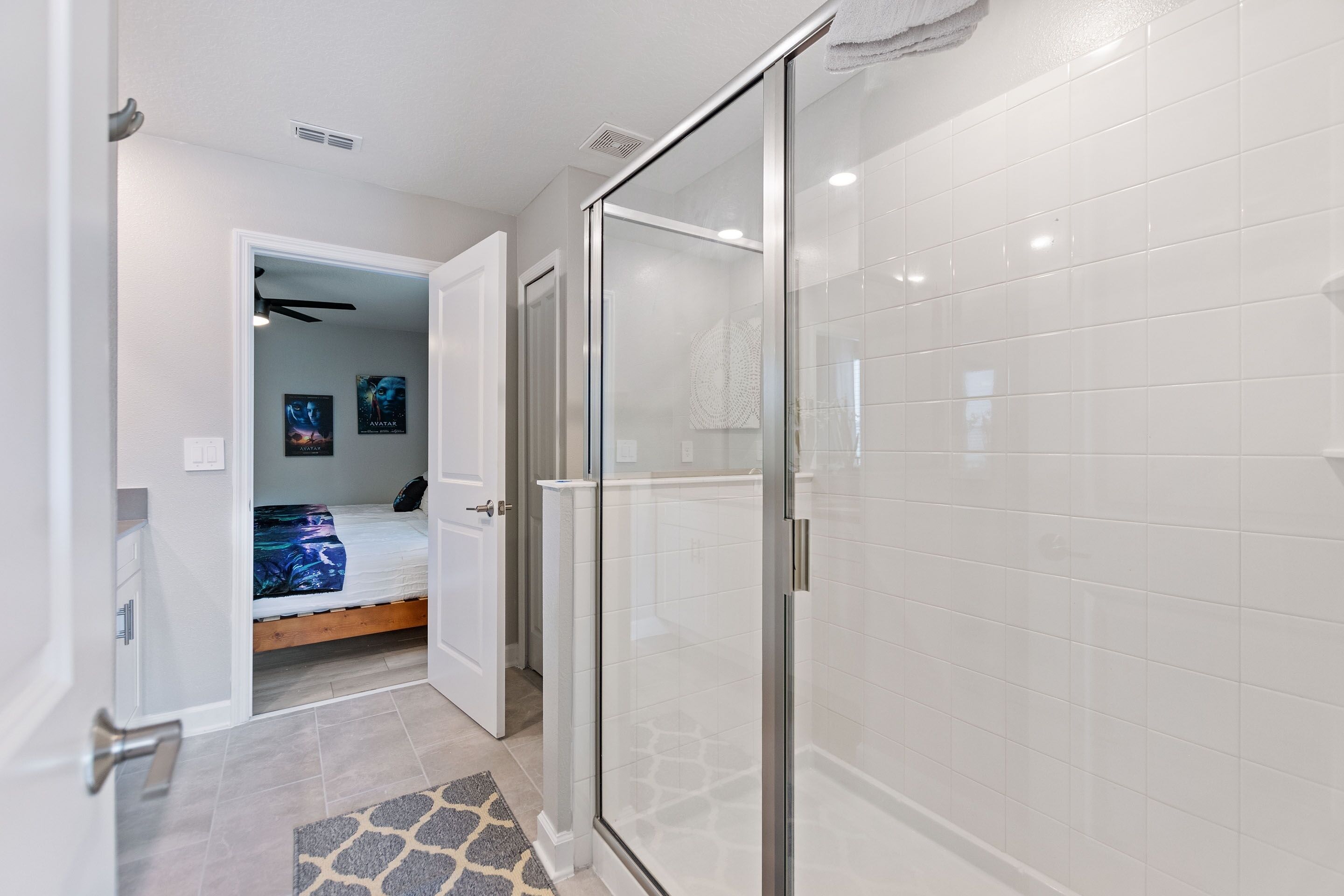 Une salle de bain moderne dotée d’une douche à parois de verre, d’un mur recouvert de carreaux blancs et d’une porte visible donnant sur une chambre à coucher avec un lit et des œuvres d’art.