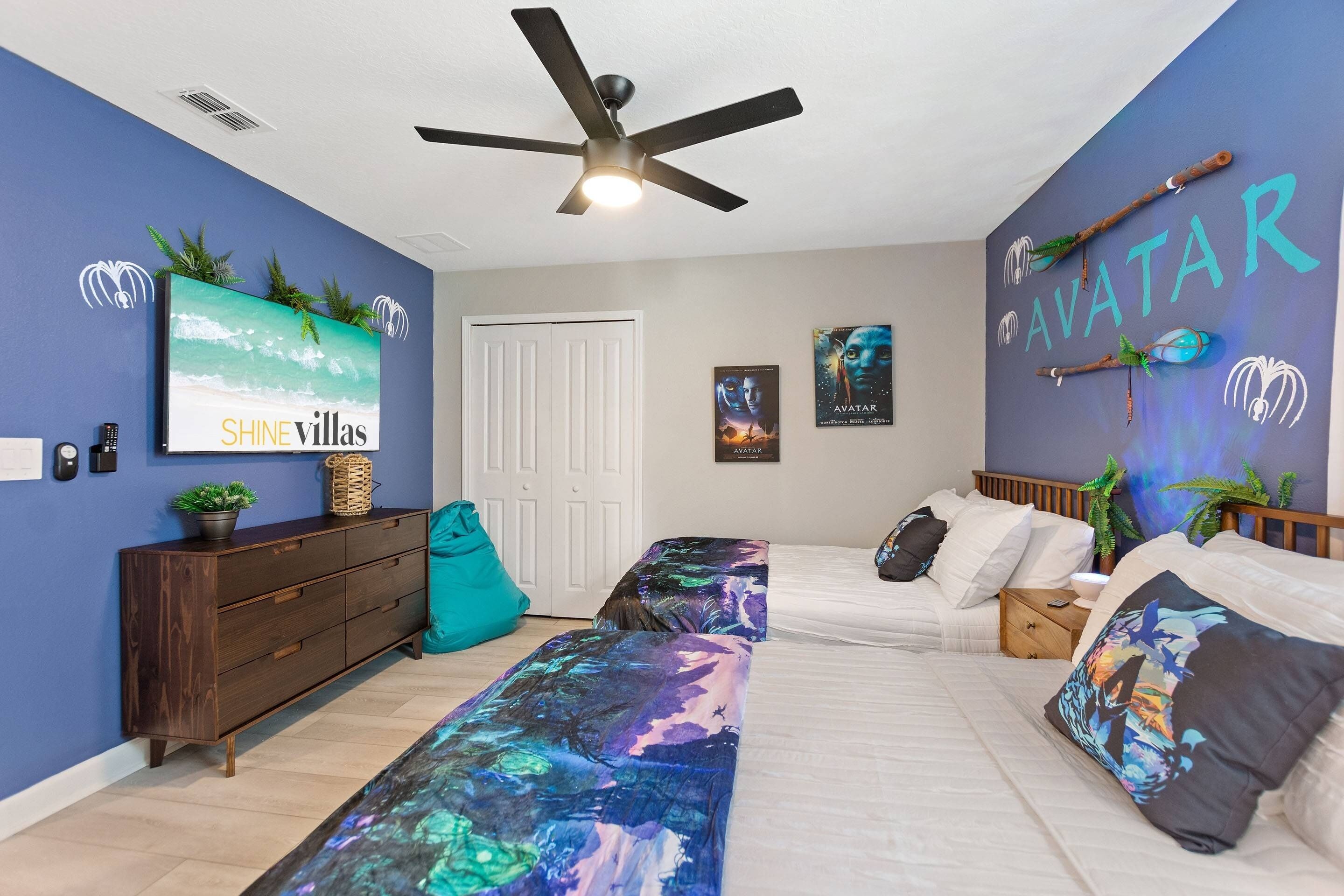 Une chambre à coucher comprenant un lit, une commode, un ventilateur de plafond et des éléments de décoration murale, notamment une affiche de « Shine Villas » et des affiches du film « Avatar ».