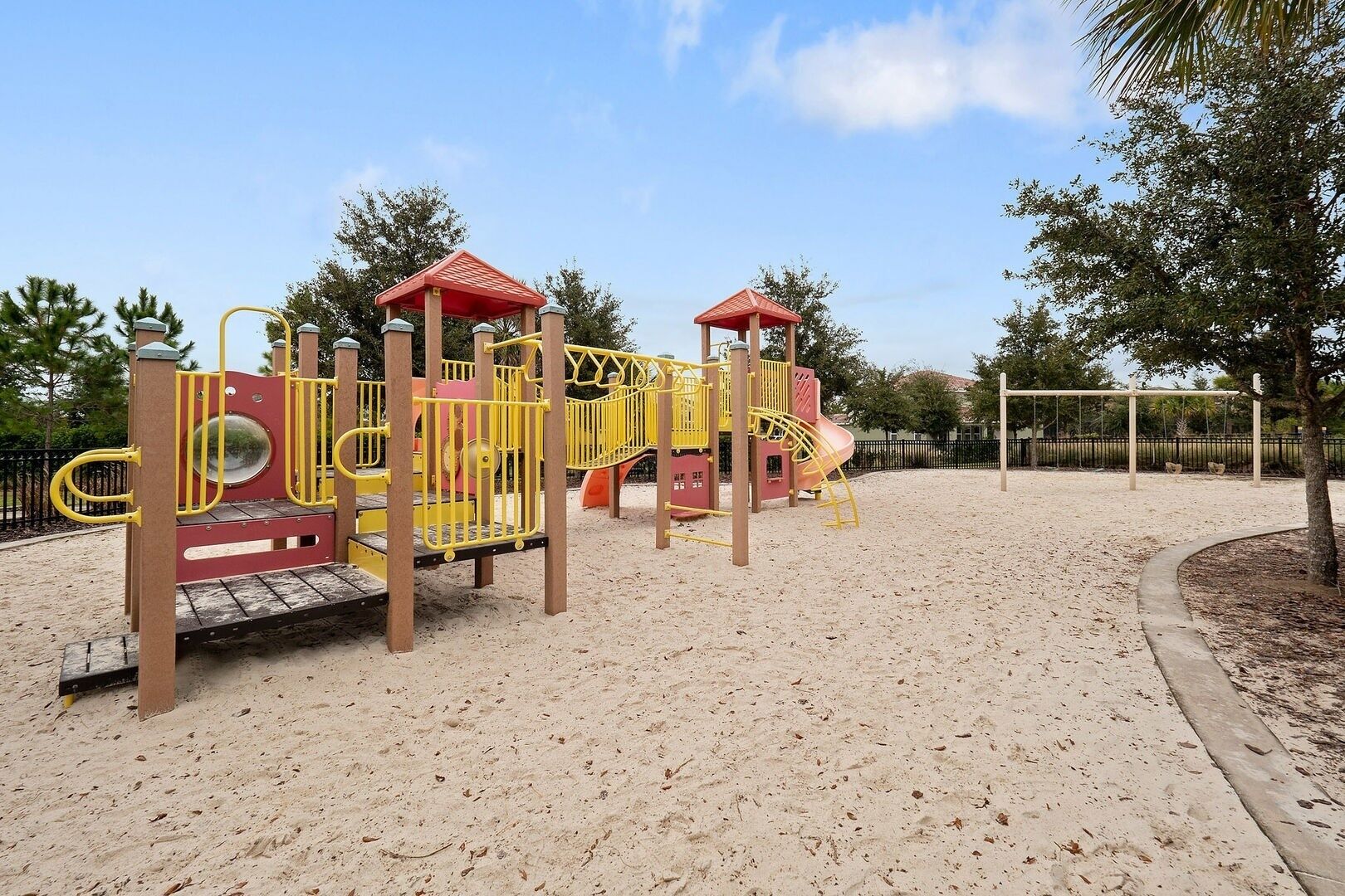 Ein Spielplatz mit Sandboden, gelben und roten Spielgeräten und einem umzäunten Bereich.