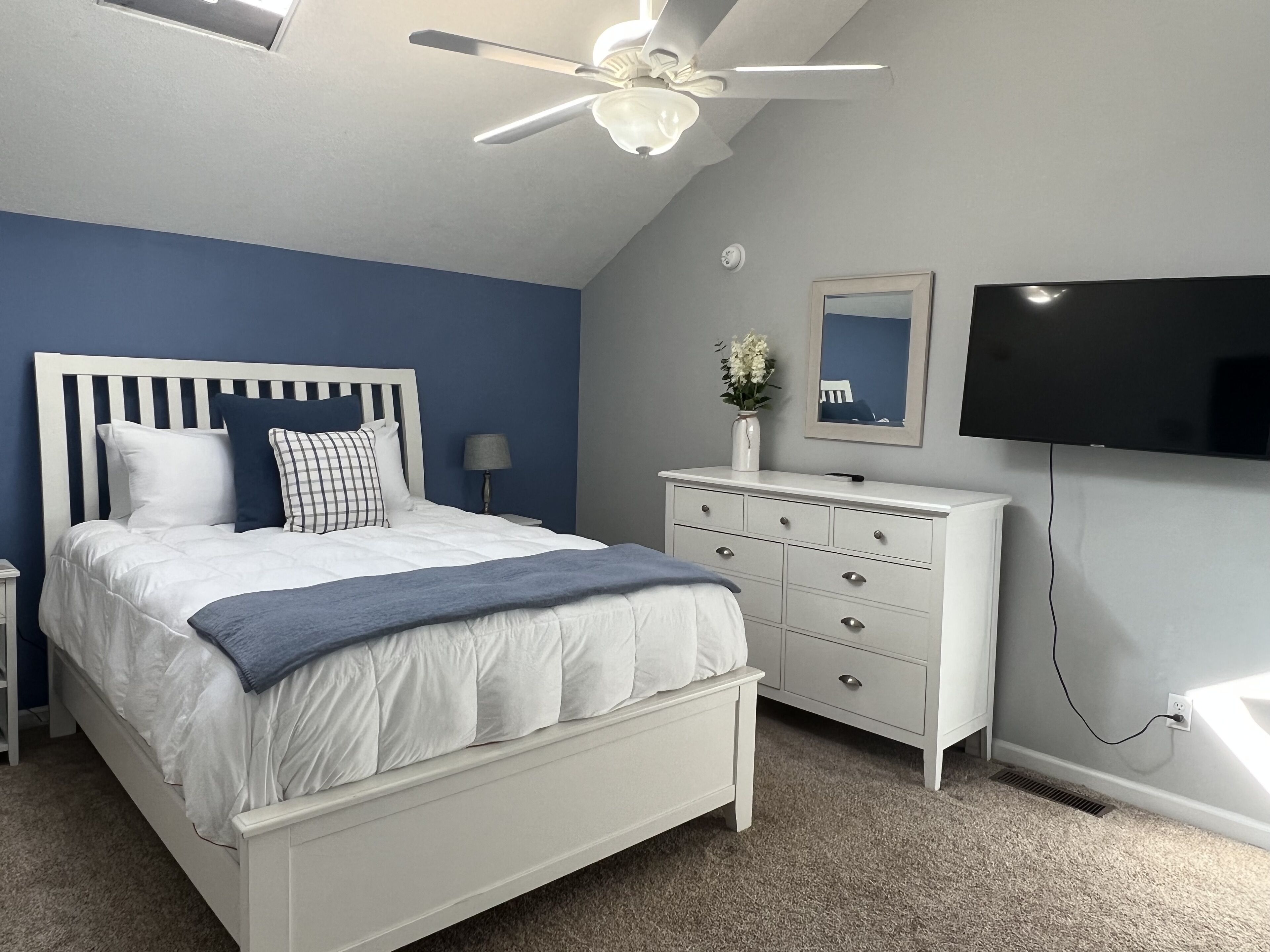 A bedroom with a bed, dresser, TV, ceiling fan, and a wall-mounted mirror.