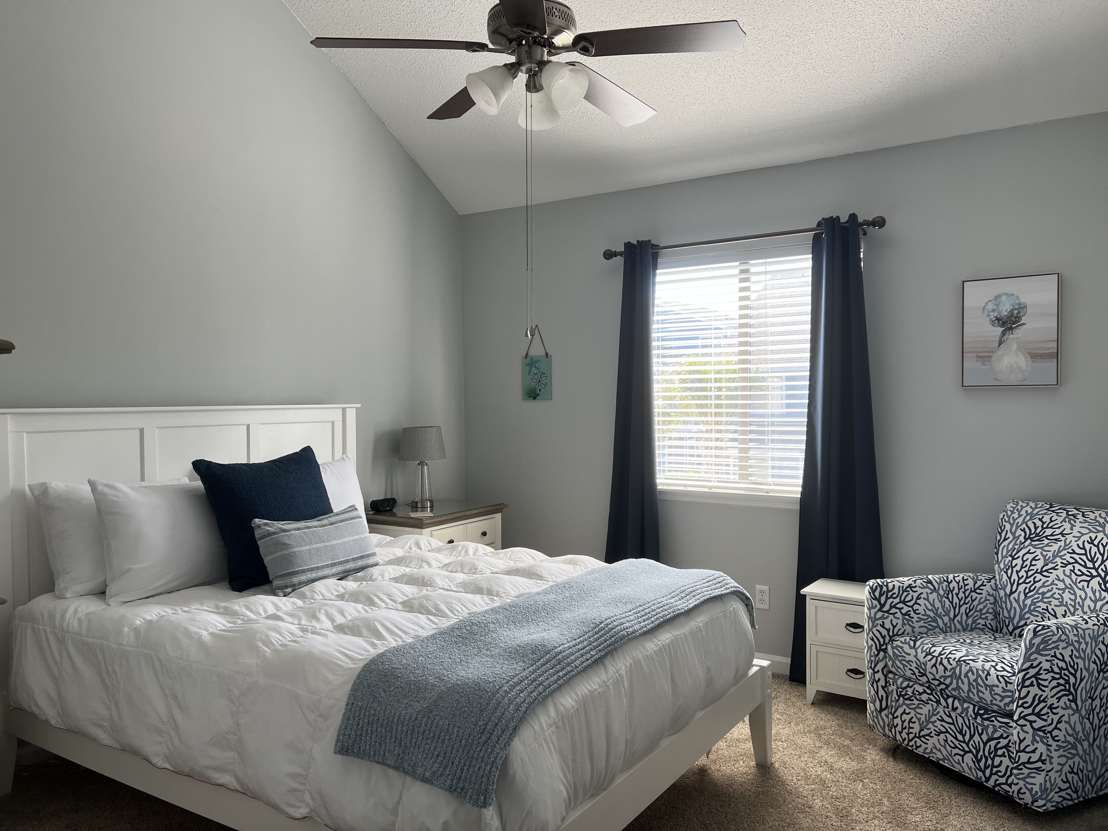 A bedroom with a bed, nightstand, chair, ceiling fan, window with curtains, and a wall-mounted artwork.