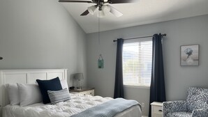 A bedroom with a bed, nightstand, chair, ceiling fan, window with curtains, and a wall-mounted artwork.