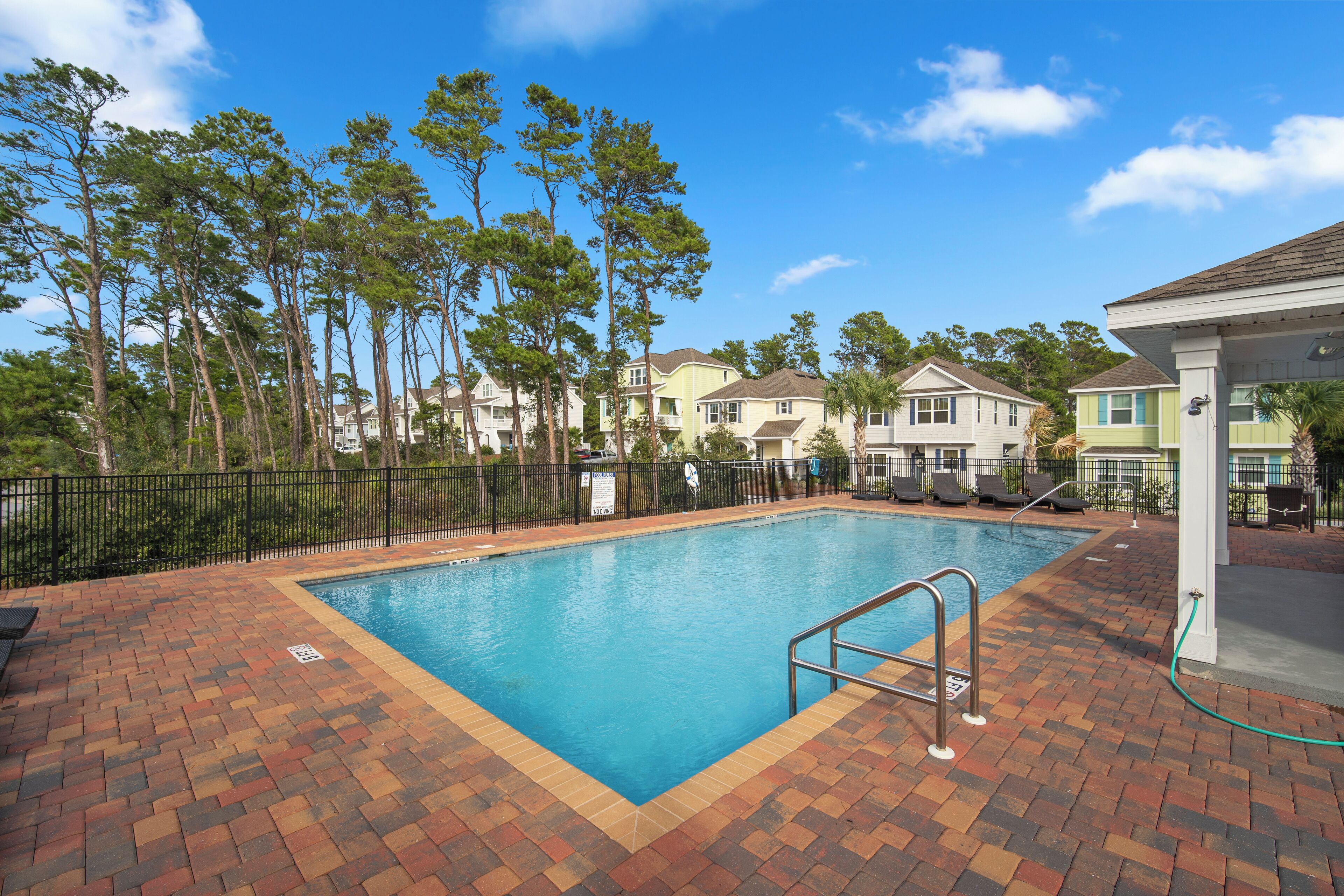 A rectangular swimming pool with a brick deck, surrounded by a black metal fence, and houses in the background.