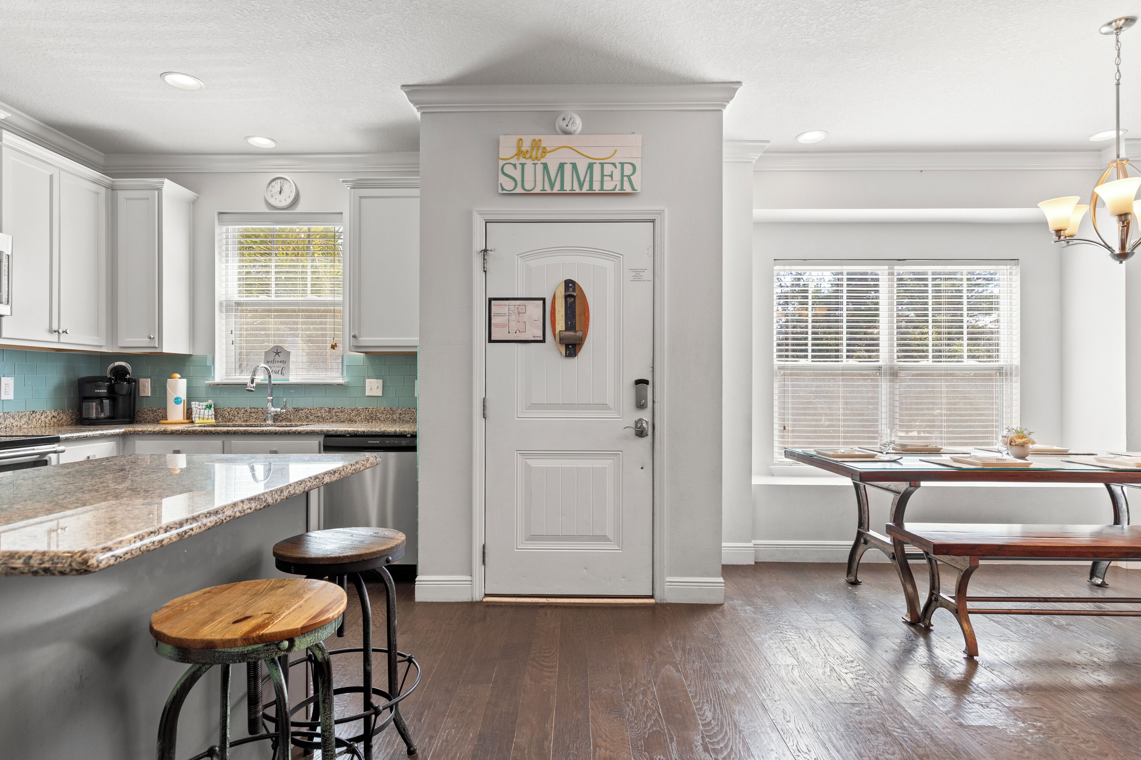 A kitchen with white cabinets, a wooden island with stools, a dining table with benches, and a 'SUMMER' sign above the door.