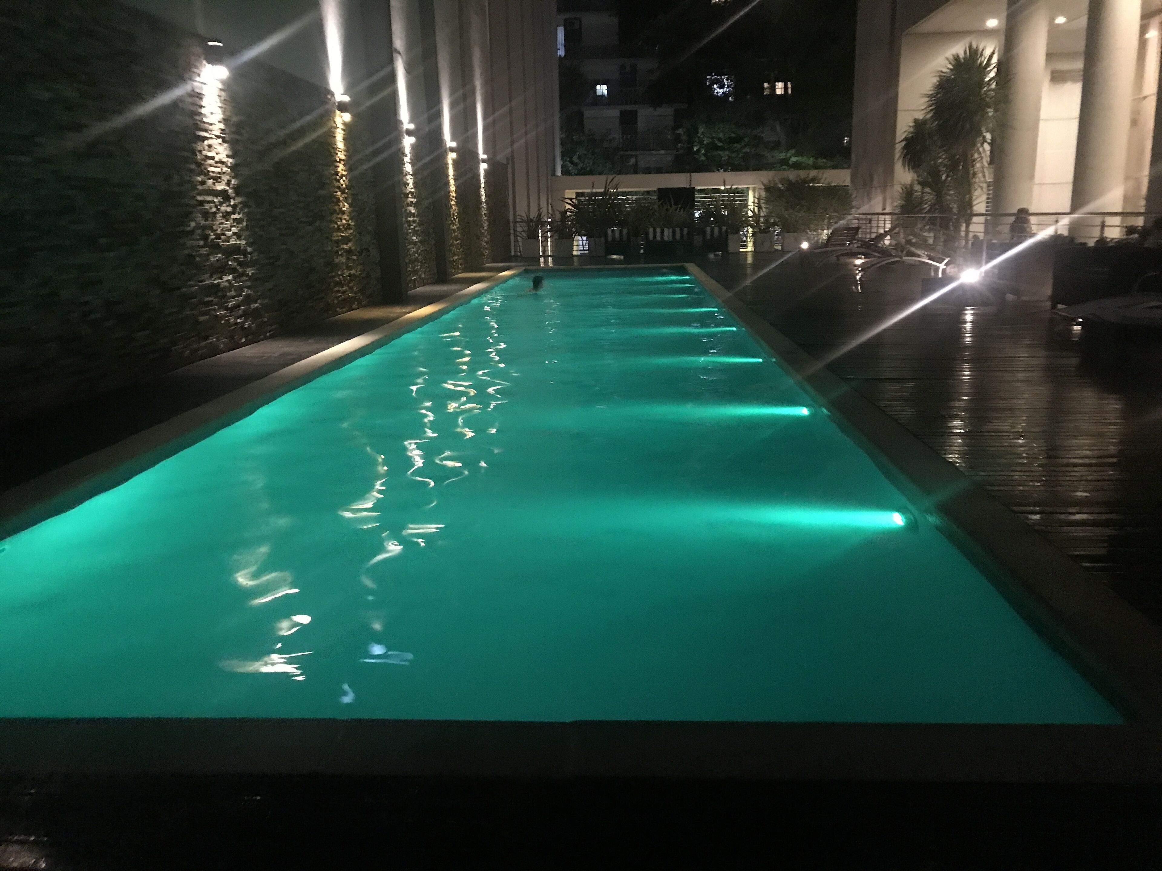 Ein beleuchteter Pool bei Nacht mit glatter Wasseroberfläche, umgeben von einer Steinmauer und Kübelpflanzen.