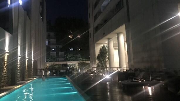 Una vista notturna di una zona piscina con una fontana, circondata da edifici con finestre illuminate e colonne.