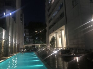 A nighttime view of a swimming pool area with a water fountain, surrounded by buildings with illuminated windows and columns.