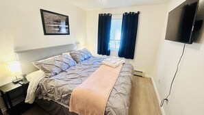 Una cama bien hecha con cabecera, una mesita de noche con lámpara, un televisor de pantalla plana montado en la pared y una ventana con cortinas.