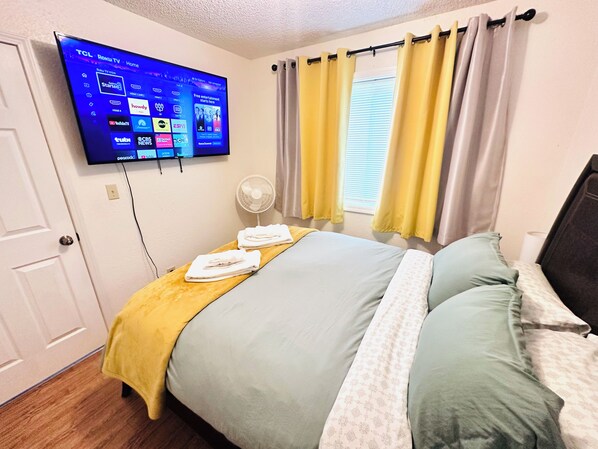 A hotel room with a bed, a flat-screen TV mounted on the wall, a window with curtains, and a fan.
