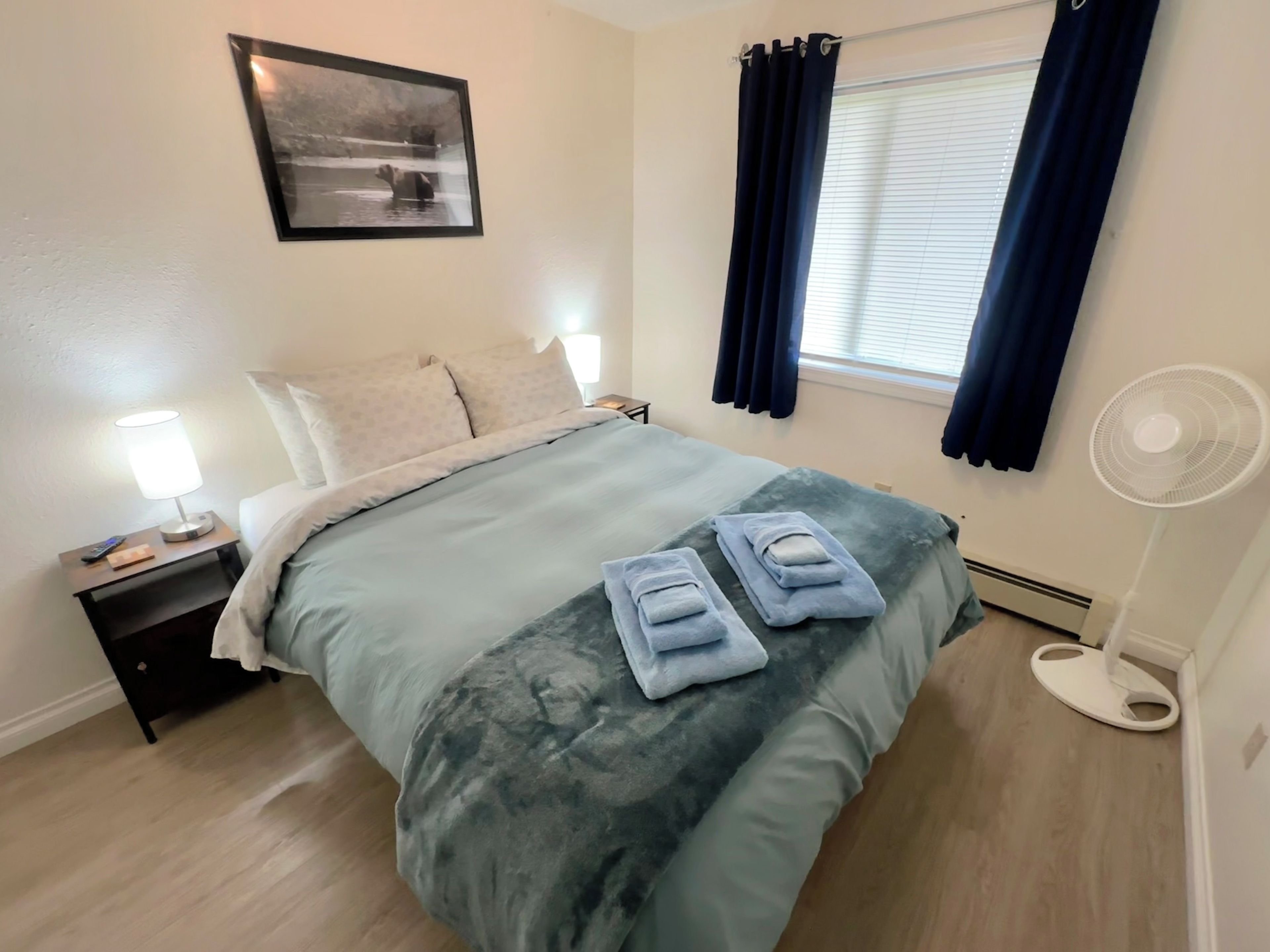 A bedroom with a bed, a nightstand, a bedside lamp, a wall-mounted picture, a window with curtains, and a standing fan.