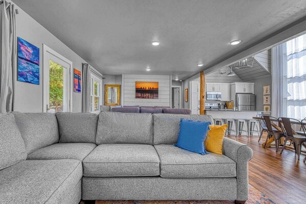 A modern living room with a grey sectional sofa, a dining area with a wooden table and chairs, and a kitchen in the background.