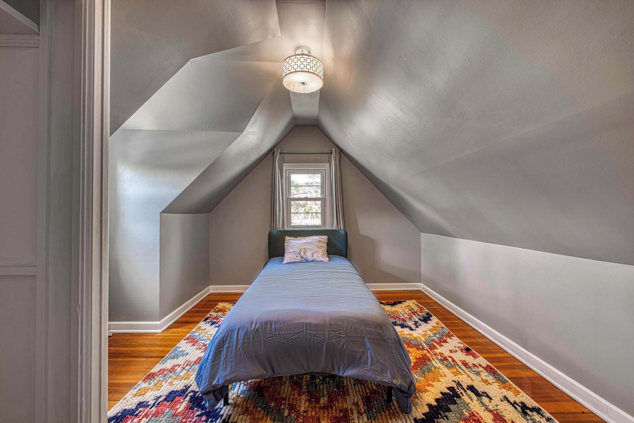 A small, sloped ceiling room with a bed, a patterned rug, and a window.
