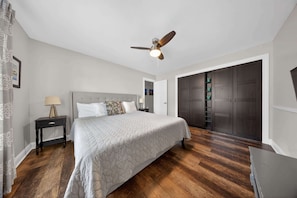 A bedroom with a bed, nightstand, lamp, ceiling fan, wardrobe, and wooden flooring. - Retro home in downtown sister bay-Sleeps 6! (Sister Bay)