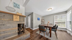 A dining area with a wooden table and chairs, a fireplace, and a view through large windows.
