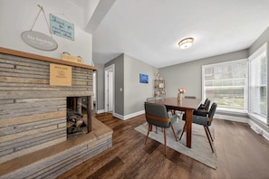 A dining area with a wooden table and chairs, a fireplace, and a view through large windows. - Retro home in downtown sister bay-Sleeps 6! (Sister Bay)