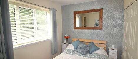 A bedroom with a bed, a nightstand, a mirror, and a window with curtains.