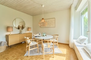 A modern living room with a round dining table, wooden chairs, a sofa, a large mirror, and a hanging light fixture.