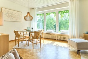 A modern living room with a wooden floor, a dining table with chairs, a sofa, a radiator, and a large window with a view of greenery. - Ferienhaus Falkenweg - Ruhig Gelegen in Einer Sackgasse mit Blick auf den Wennsee in Scharbeutz. - (Scharbeutz)