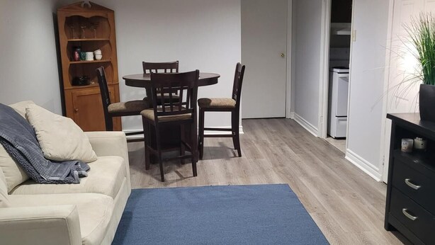 A living room with a white sofa, a blue rug, a wooden dining table with chairs, a bookshelf, and a kitchen area visible through an open door. - Cozy resting place for one (Toronto)