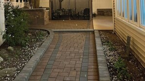 Un área de estar al aire libre cubierta con luces colgantes, un camino de ladrillo y una valla de madera.